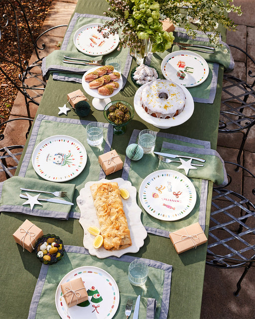 A festive outdoor table for four with green linens, Idda Studio x Bed Threads 'Caramella' ceramic dinner plates, gift-wrapped boxes, a bundt cake, cookies on a handmade plate, snacks, and a vase of fresh greenery.