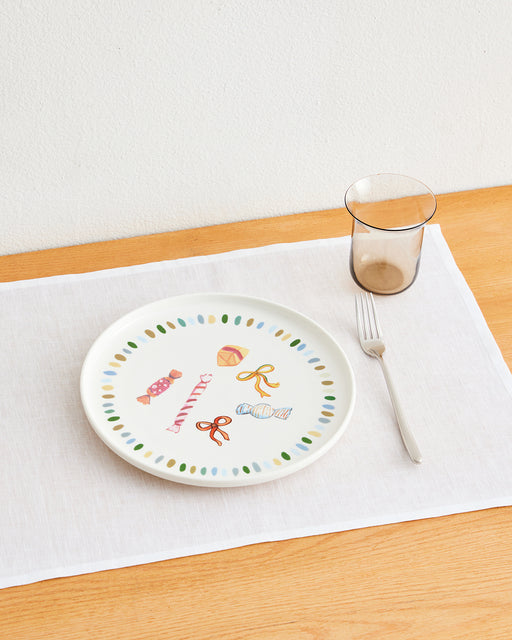 The Idda Studio x Bed Threads 'Caramella' Ceramic Dinner Plate, featuring colorful candy designs, rests on a white placemat beside a fork and a translucent brown glass atop a light wooden table.