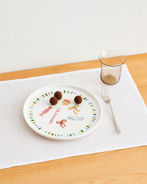 An Idda Studio x Bed Threads 'Caramella' Ceramic Dinner Plate holds four chocolate truffles, placed on a white placemat with a fork and clear glass atop a light wooden table.