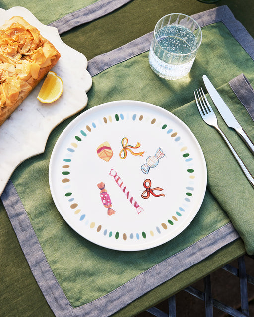 An Idda Studio x Bed Threads 'Caramella' Ceramic Dinner Plate decorated with colorful candy motifs rests on a green and gray placemat beside cutlery, a glass of water, and a marble tray with a pastry and lemon wedge.