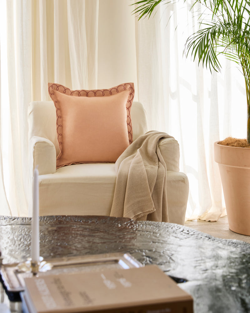 A cream armchair with a Bed Threads Terracotta & Pink Clay French Flax Linen Swirl Cushion Cover and beige throw sits by a large potted plant near sheer curtains. A black table with a book is in front, bathed in soft, natural light.