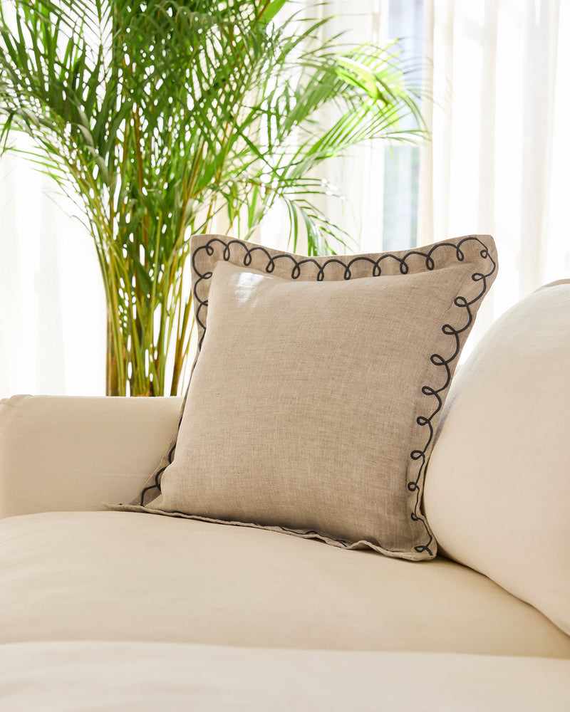 A Bed Threads Oatmeal & Charcoal 100% French Flax Linen Swirl Cushion Cover with decorative dark trim rests on a cream sofa, with sunlight shining through sheer curtains and a tall green plant in the background.