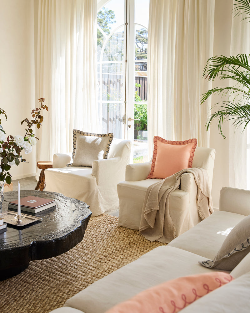 Bright, airy living room with cream armchairs and sofa, pastel throw pillows, Bed Threads’ Oatmeal 100% French Flax Linen Muslin Throw, large windows with sheer curtains, black round coffee table, potted plant, and woven rug for a cozy atmosphere.