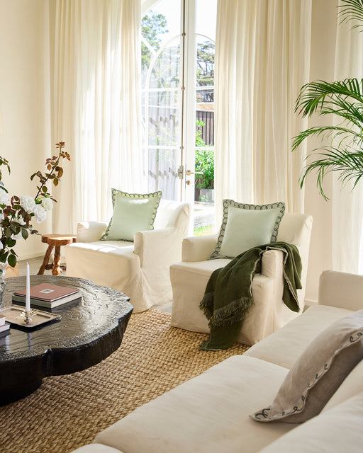 Bright living room with white armchairs, green cushions, a black coffee table, woven rug, potted plant, and Bed Threads' Oatmeal & Olive 100% French Flax Linen Muslin Throw for textured accents. Sheer curtains welcome natural light in this serene space.