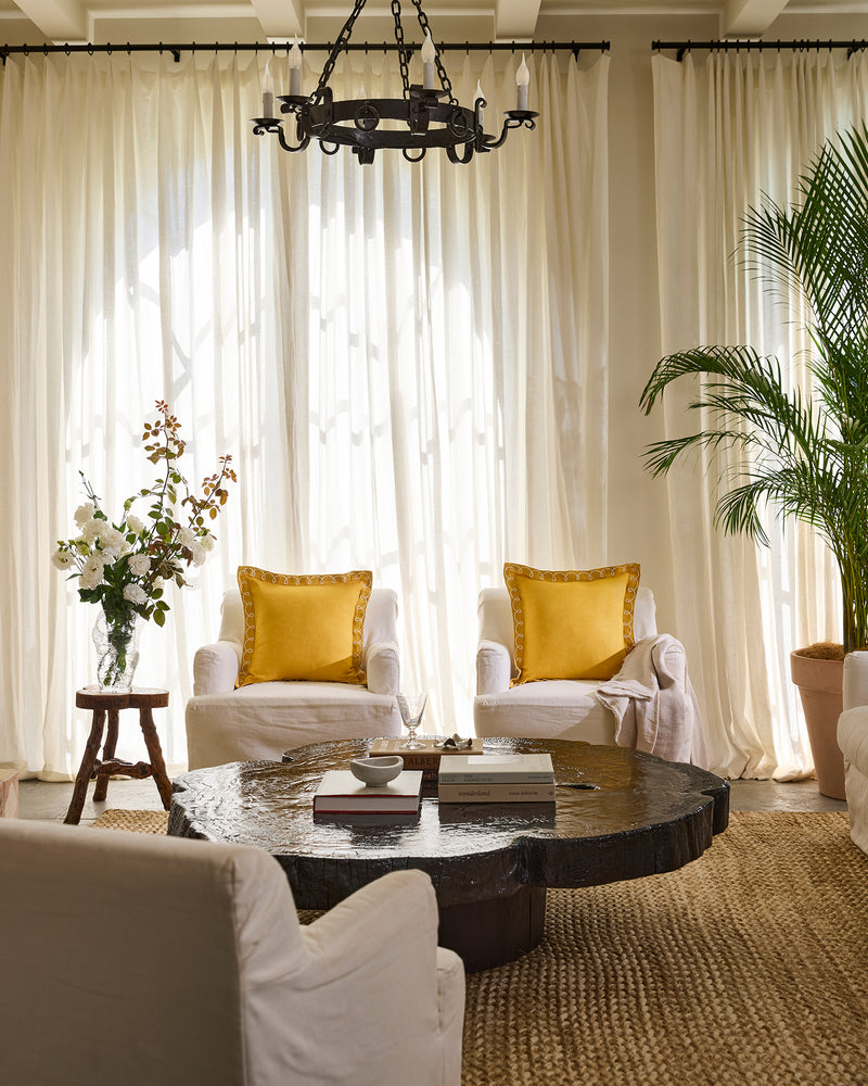 A cozy living room with two white armchairs, each featuring a yellow pillow and a Bed Threads Turmeric & Crème 100% French Flax Linen Swirl Cushion Cover, rustic round wooden coffee table, potted plants, and sheer white curtains with natural light.