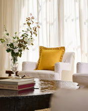 A cozy living room with a white armchair topped by Bed Threads’ Turmeric & Crème 100% French Flax Linen Swirl Cushion Cover, a glass vase of white and brown flowers on a side table, books on a dark coffee table, and sheer white curtains. - view