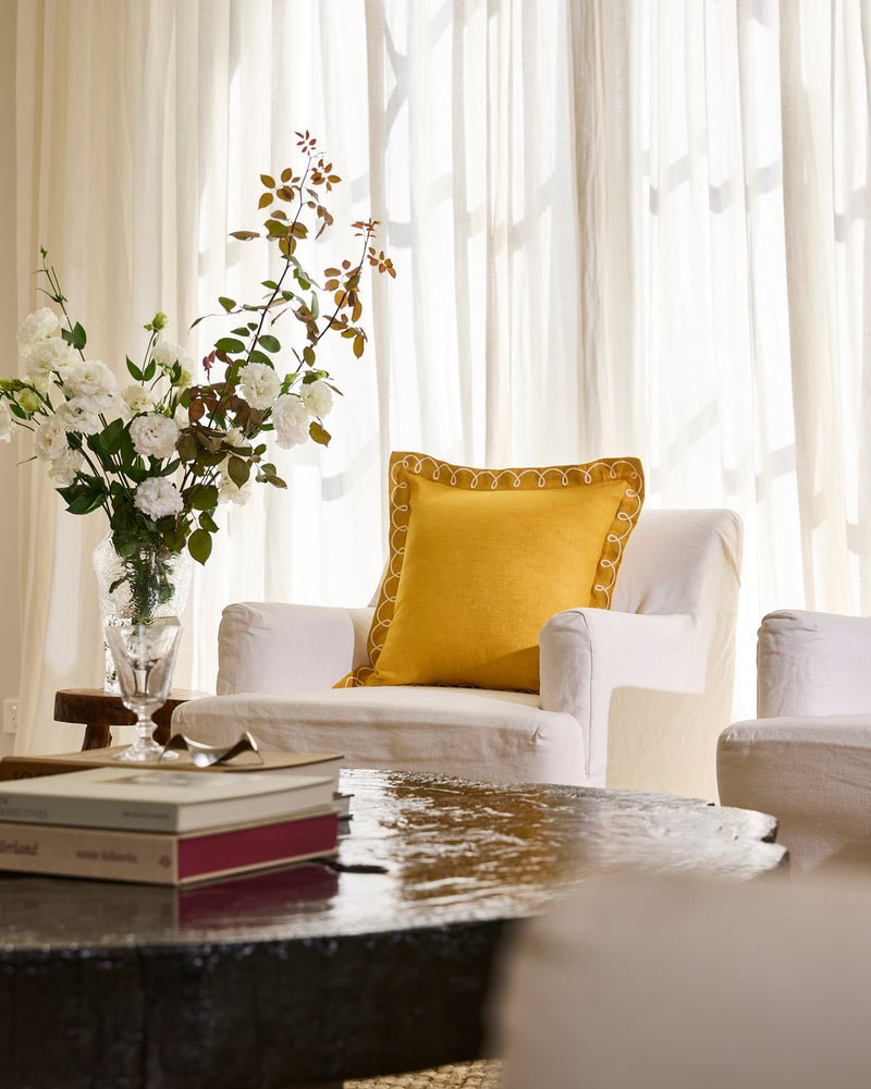 A cozy living room with a white armchair topped by Bed Threads’ Turmeric & Crème 100% French Flax Linen Swirl Cushion Cover, a glass vase of white and brown flowers on a side table, books on a dark coffee table, and sheer white curtains.