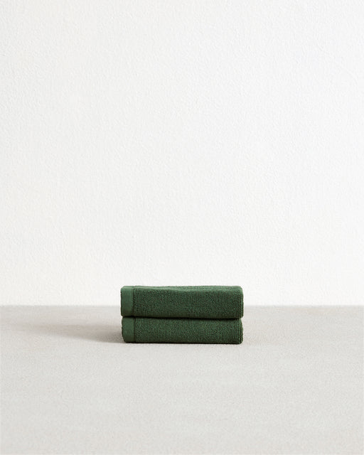 The Bed Threads Moss 100% Cotton Terry Face Washers (Set of Two) are soft, absorbent dark green towels, neatly folded on a light grey surface against a white wall—ideal for daily use.