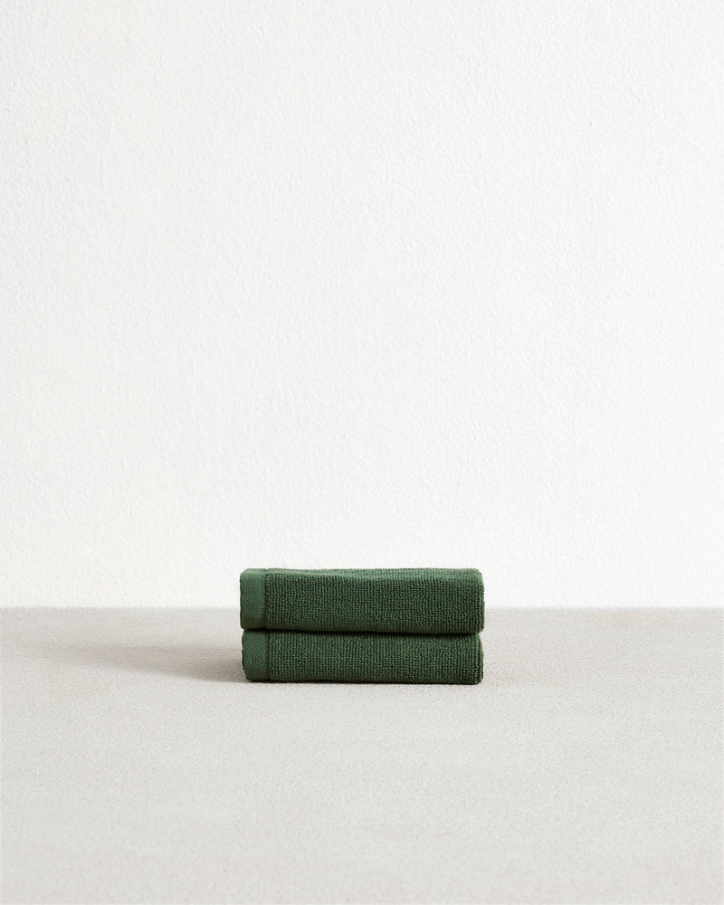 The Bed Threads Moss 100% Cotton Terry Face Washers (Set of Two) are soft, absorbent dark green towels, neatly folded on a light grey surface against a white wall—ideal for daily use.