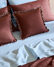 Two maroon decorative pillows with white scalloped edges and a Cacao & Coast 100% French Flax Linen Arch Cushion Cover by Bed Threads rest on light blue sheets and maroon pillowcases, creating a cozy, contrasting look. - view
