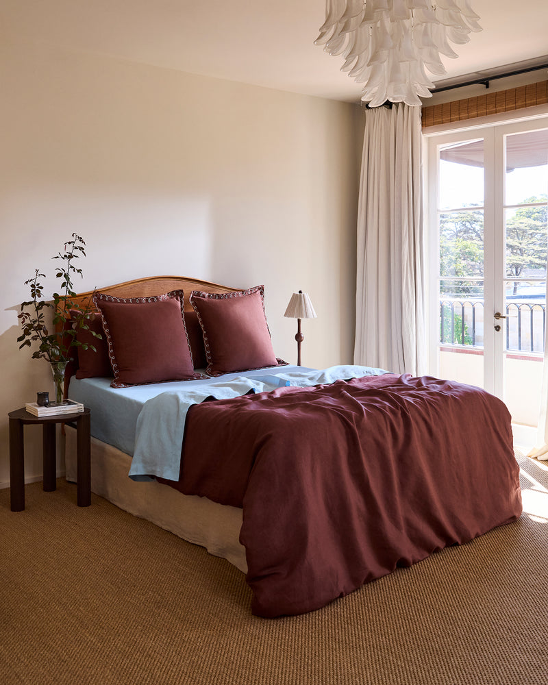 A cozy bedroom with a neatly made bed featuring Bed Threads' Cacao & Coast 100% French Flax Linen Swirl European Pillowcases, a blue sheet, a bedside table with a plant and books, a floor lamp, and large windows adorned with white curtains.