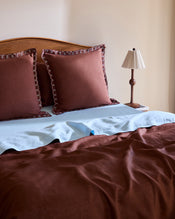 A neatly made bed with Bed Threads Cacao & Coast 100% French Flax Linen Swirl European Pillowcases, light blue French flax linen sheets (170GSM), a brown bedspread, and a small bedside table holding a brown lamp and book. - view