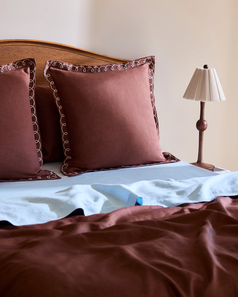 A neatly made bed features Bed Threads’ Cacao & Coast 100% French Flax Linen Swirl European Pillowcases (Set of Two) with decorative edging, a light blue linen sheet, a brown blanket, and a small lamp on the wooden nightstand.