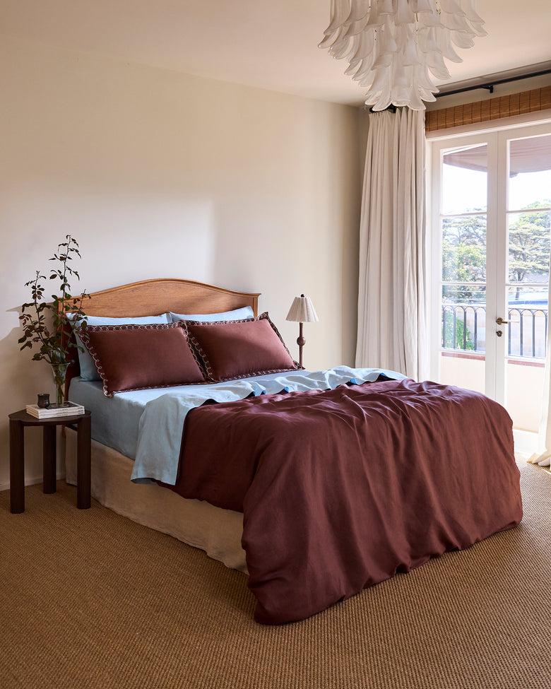 A neatly made bed with burgundy bedding and light blue sheets stands beside a small wooden table with books. Theres a potted plant, a standing lamp, and large windows with white curtains letting in natural light.