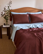 A neatly made bed with a wooden headboard features light blue sheets and brown bedding, topped with Bed Threads' Cacao & Coast 100% French Flax Linen Swirl Pillowcases. Nearby, a round table holds a vase with branches and a stack of books. - view