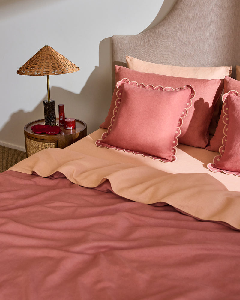 A neatly made bed with rose and blush bedding, scalloped-edge pillows, and a headboard is accented by the Bed Threads Pink Clay & Terracotta 100% French Flax Linen Arch Cushion Cover. A round side table holds a wicker lamp, candle, and red bottle for a cozy touch.