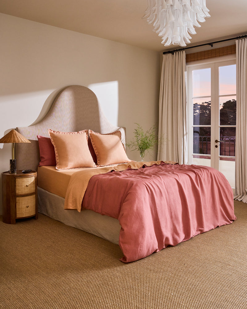 A cozy bedroom features a uniquely shaped upholstered headboard, Bed Threads’ Terracotta & Pink Clay 100% French Flax Linen Swirl European Pillowcases, a wooden nightstand with a lamp, sheer curtains, a glass door, and a modern chandelier.