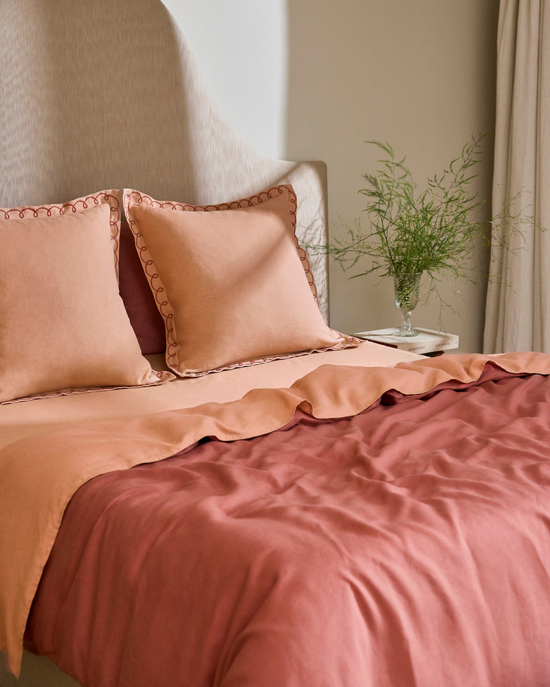 A bed styled with Bed Threads Terracotta & Pink Clay 100% French Flax Linen Swirl European Pillowcases, a beige upholstered headboard, and a side table topped with a glass vase of green foliage.