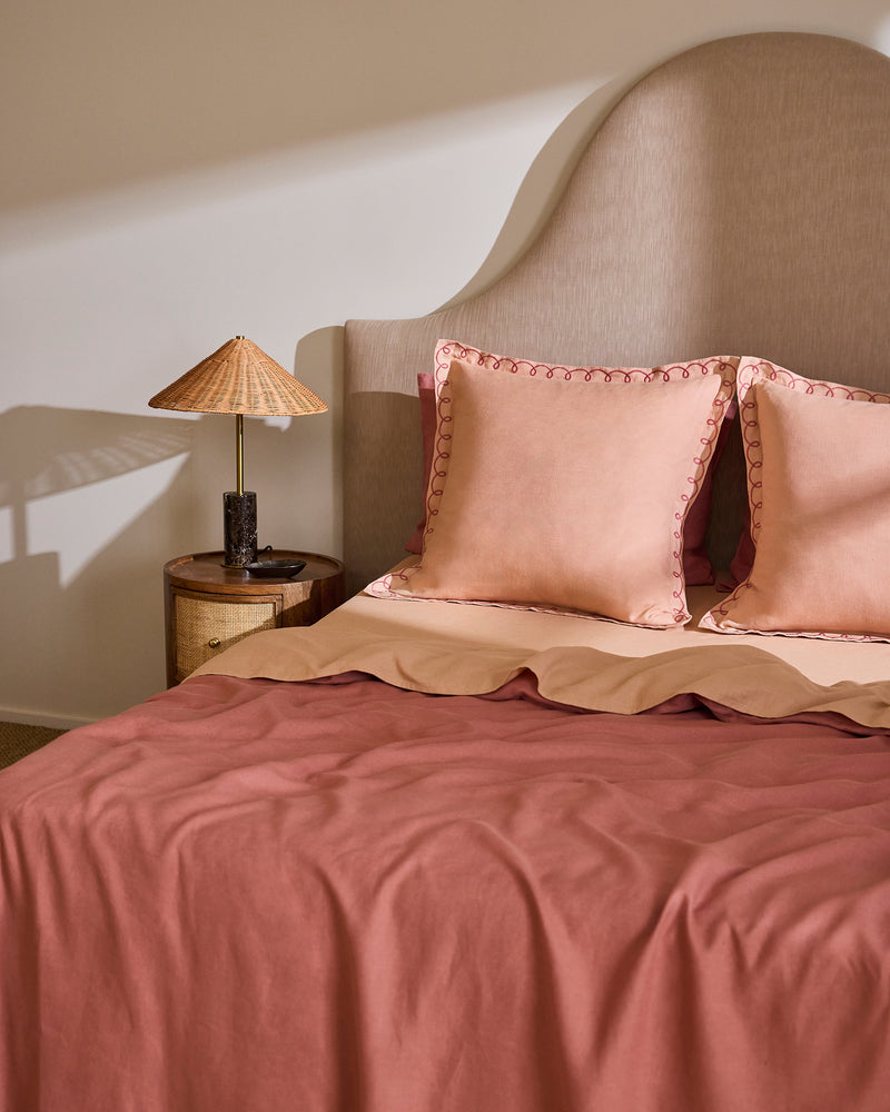 A neatly made bed features Bed Threads’ Terracotta & Pink Clay 100% French Flax Linen Swirl European Pillowcases (Set of Two), beside a small round wooden nightstand with a stone-based lamp and woven lampshade against a light wall.