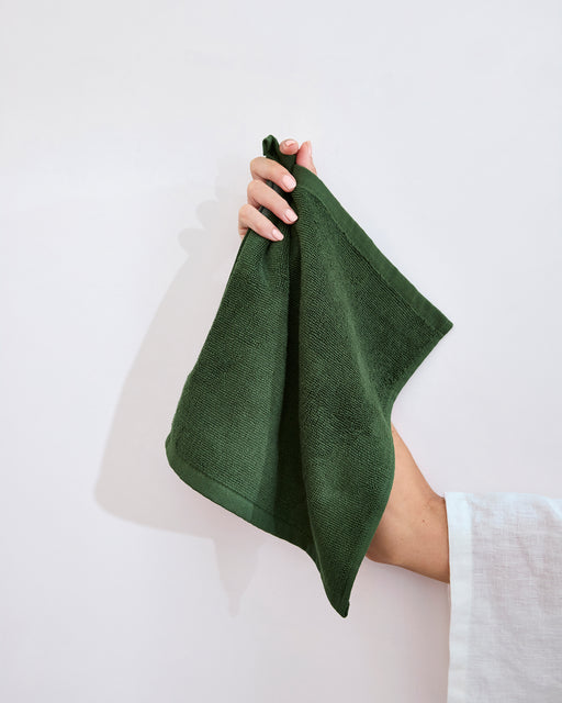 A hand with a light sleeve holds up a Moss 100% Cotton Terry Face Washer by Bed Threads, showcasing its soft, absorbent texture—ideal for daily care—against a plain light background. Sold as a set of two.