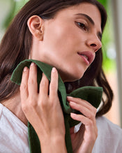 A woman with long brown hair, wearing a white top and a calm expression, gently presses a soft Moss 100% Cotton Terry Face Washer from Bed Threads (Set of Two) to her neck. - view