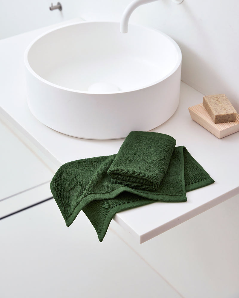 A modern white bathroom sink features a folded dark green towel and Bed Threads Moss 100% Cotton Terry Face Washers (Set of Two) on the counter, with a beige sponge in a wooden dish creating an inviting daily care space.
