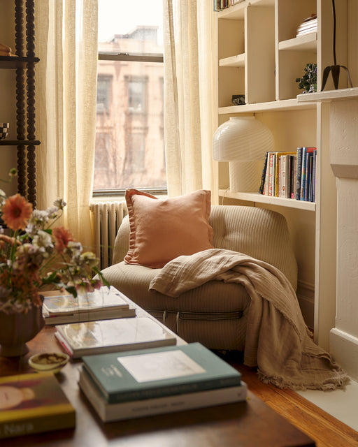 A cozy reading nook features a beige armchair with a peach pillow and the Bed Threads Terracotta 100% French Flax Linen Cushion Cover by the window, sheer curtains, shelves of books, a lamp, and fresh flowers creating a warm, inviting atmosphere.