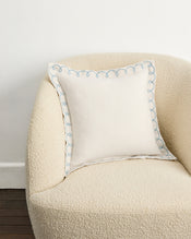 A cream armchair with a Bed Threads Crème & Coast 100% French Flax Linen Swirl Cushion Cover, featuring light blue embroidery, sits against a white wall and dark wood floor. - view
