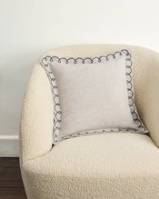 A cream textured armchair with a swirl edge is paired with the Bed Threads Oatmeal & Charcoal 100% French Flax Linen Swirl Cushion Cover, showcasing a black embroidered border, against a white wall and dark wood floor. - view
