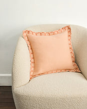 A soft, cream-colored textured armchair with a Bed Threads Terracotta & Pink Clay 100% French Flax Linen Swirl Cushion Cover sits against a plain white wall and dark wooden floor. - view