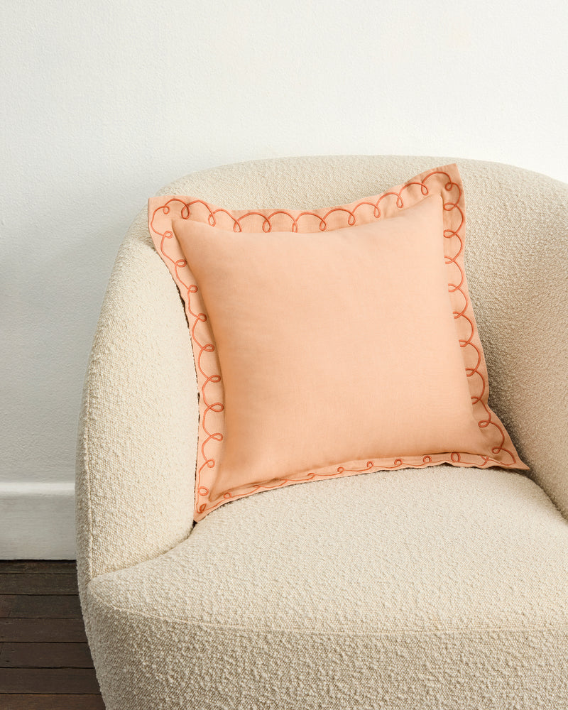 A soft, cream-colored textured armchair with a Bed Threads Terracotta & Pink Clay 100% French Flax Linen Swirl Cushion Cover sits against a plain white wall and dark wooden floor.