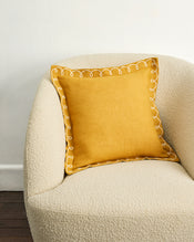 A Turmeric & Crème 100% French Flax Linen Swirl Cushion Cover by Bed Threads sits on a cream-colored textured armchair against a plain white wall. - view