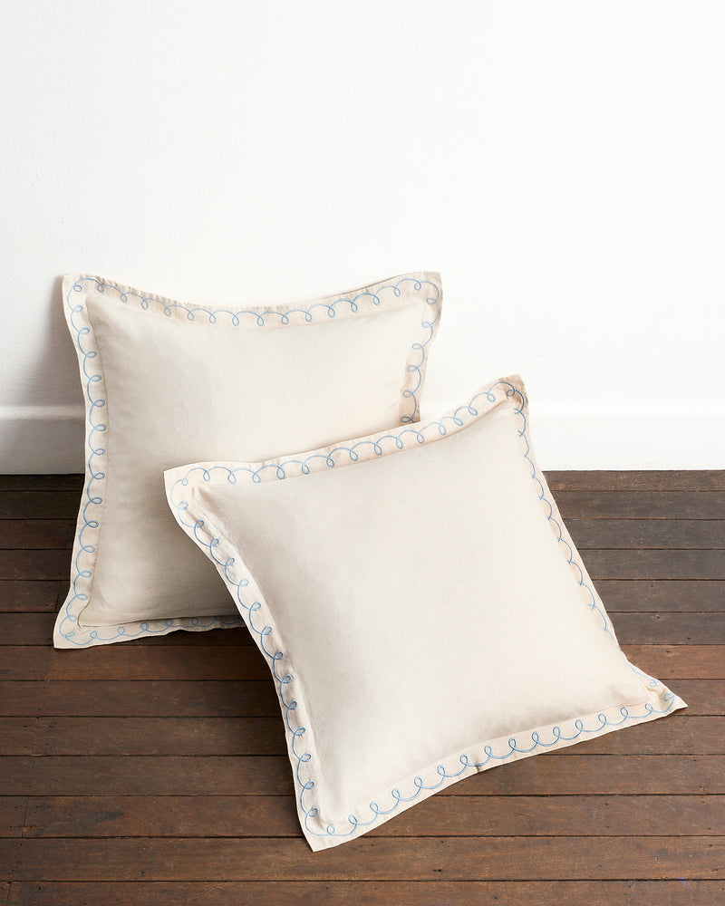 Two Crème & Coast 100% French Flax Linen Swirl European Pillowcases by Bed Threads, set of two, in beige with light blue decorative edge stitching and zip closure, displayed on a wooden floor against a white wall.