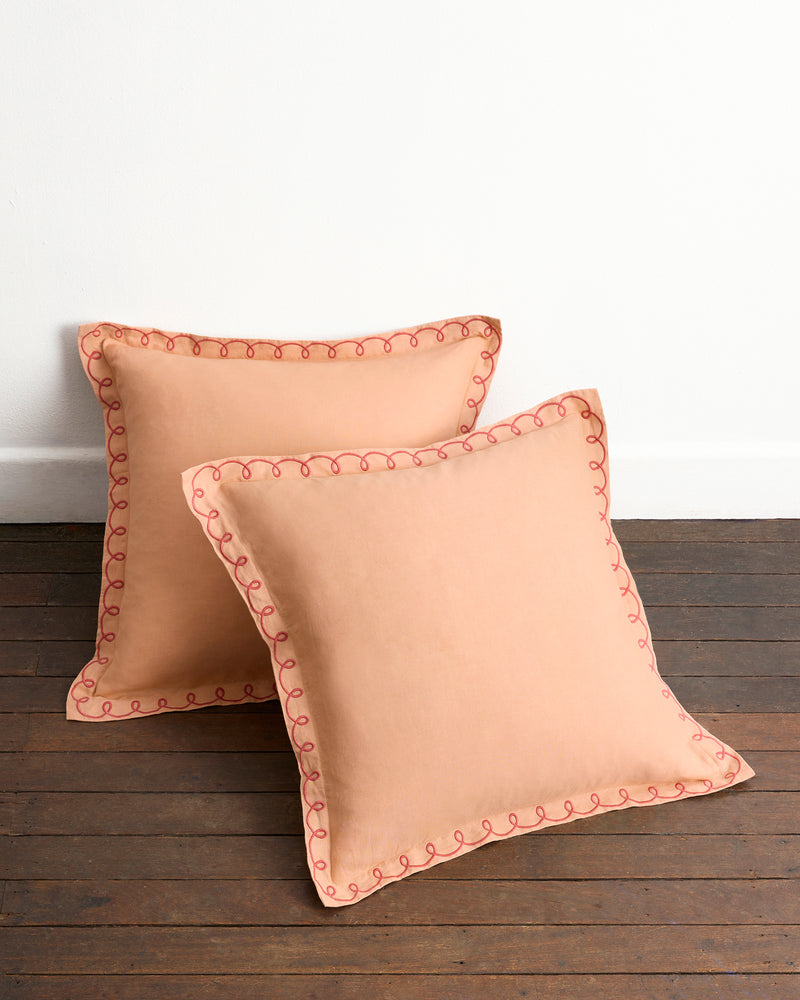 The Bed Threads Terracotta & Pink Clay 100% French Flax Linen Swirl European Pillowcases (Set of Two) are displayed on a wooden floor, leaning against a white wall, highlighting their pink embroidered trim and hidden zip closure.
