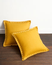 Two Turmeric & Crème 100% French Flax Linen Swirl European Pillowcases by Bed Threads are arranged on a wooden floor against a white wall—one upright, the other leaning—in soft yellow with white embroidered trim. - view