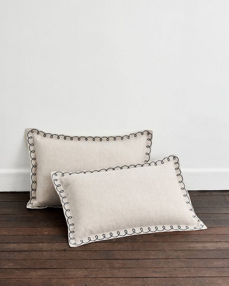 A pair of Bed Threads Oatmeal & Charcoal 100% French Flax Linen Swirl Pillowcases rests on a dark wooden floor against a plain white wall.