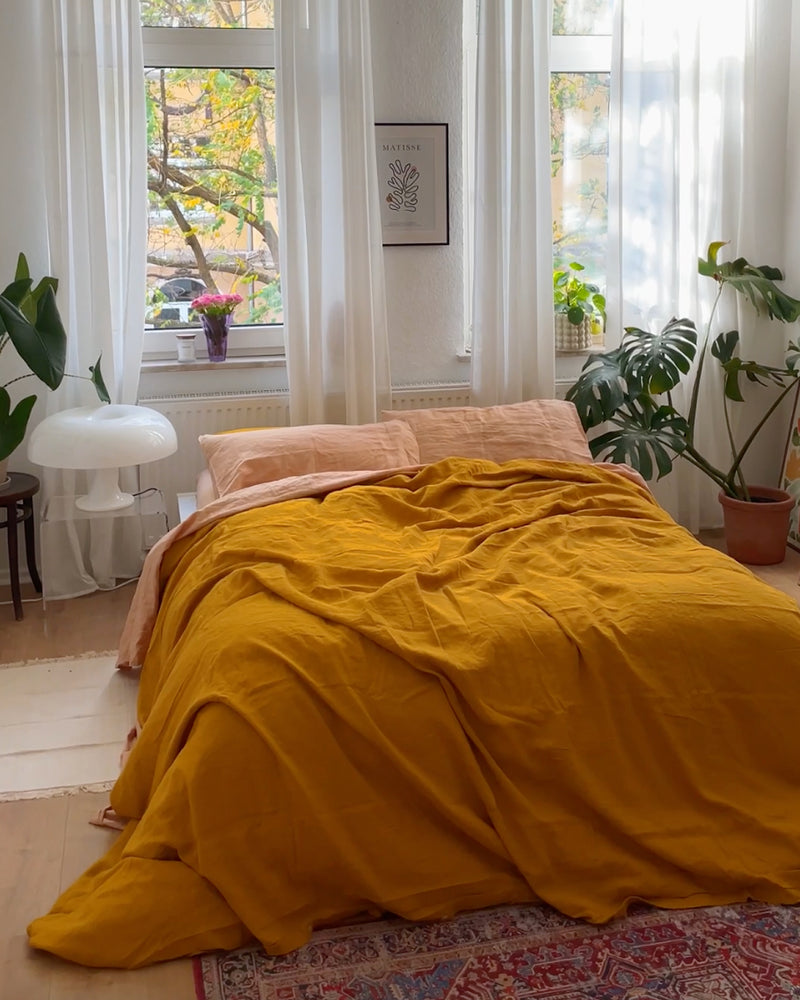 A cozy bedroom styled with the Bed Threads Turmeric & Terracotta 100% French Flax Linen Bedding Bundle, pale pink pillows, potted plants, large sunlit windows, a white lamp, and a patterned rug on the floor.
