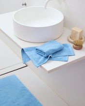 A modern white bathroom sink with a round basin features a bar of soap, scrubbing brush, and neatly folded Bed Threads Sky 100% Cotton Terry Face Washers (Set of Two) for daily care. A matching absorbent terry blue bath mat is on the floor. - view