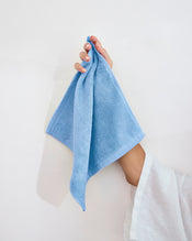 A hand holds a Sky 100% Cotton Terry Face Washer from Bed Threads (Set of Two) against a plain white background. The person, in a white long-sleeve top, shows how these absorbent washers suit daily care routines. - view