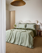A cozy bedroom features a wavy cream headboard, sage green bedding, and Bed Threads' Sage & Olive 100% French Flax Linen Swirl Pillowcases. Two pillows, a wooden side table with white lamp, woven pendant, natural fiber carpet, and flowers complete the look. - view