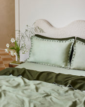A neatly made bed features Bed Threads’ Sage & Olive 100% French Flax Linen Swirl Pillowcases, an olive green blanket, and a light green duvet. A vase with white flowers rests on the bedside table next to the bed. - view