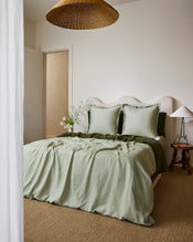 A cozy bedroom with a bed made up in sage green French flax linen, two Bed Threads Sage & Olive 100% French Flax Linen Swirl European Pillowcases, a textured beige headboard, wooden side table and lamp, wicker pendant light, and neutral décor. - view