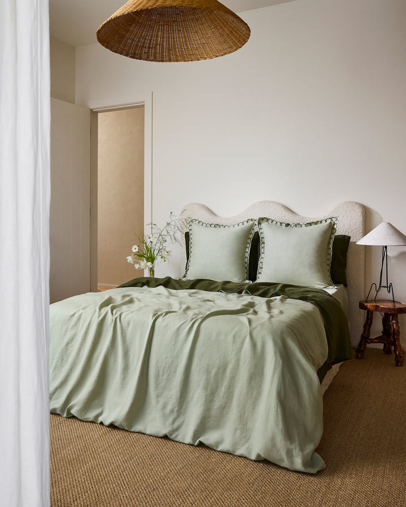 A cozy bedroom with a bed made up in sage green French flax linen, two Bed Threads Sage & Olive 100% French Flax Linen Swirl European Pillowcases, a textured beige headboard, wooden side table and lamp, wicker pendant light, and neutral décor.