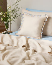 A neatly made bed with Bed Threads' Crème & Coast 100% French Flax Linen Arch Cushion Cover, light blue sheets, a matching cream blanket, and a glass vase with white flowers and greenery on the bedside table. - view