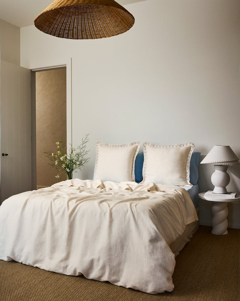 A cozy bedroom with a neatly made bed featuring Bed Threads' Crème & Coast 100% French Flax Linen Swirl European Pillowcases (Set of Two), a white sculpted lamp on a side table, woven pendant light, and minimal decor including greenery in a vase.