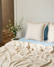 A neatly made bed features Bed Threads' Crème & Coast 100% French Flax Linen Swirl European Pillowcases (Set of Two) in cream, set against a light wall and beside a small table with a vase of white flowers and greenery. - view