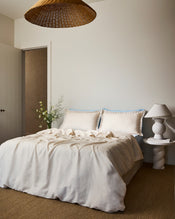 A cozy bedroom with a neatly made bed featuring Bed Threads' Crème & Coast 100% French Flax Linen Swirl Pillowcases (Set of Two), a small white side table with a decorative lamp, a flower vase, and a woven pendant light overhead. - view