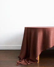 A wooden table draped with the Bed Threads Cacao 100% French Flax Linen Tablecloth sits on a dark wood floor against a plain white wall. - view