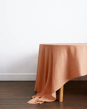 A round wooden dining table, partially draped with the Hazelnut 100% French Flax Linen Tablecloth by Bed Threads, sits on a wooden floor against a plain white wall. - view