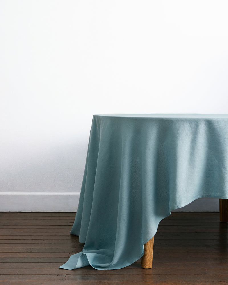 The Lagoon 100% French Flax Linen Tablecloth by Bed Threads in light blue elegantly drapes over a wooden table, with one corner cascading toward the floor against a white wall, adding sophistication to your dining space.